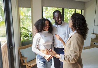 A man and woman smile as they speak with an apartment employee during a tour of a potential new home, with large open windows providing a bright, welcoming view in the background.