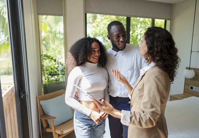 A man and woman smile as they speak with an apartment employee during a tour of a potential new home, with large open windows providing a bright, welcoming view in the background.