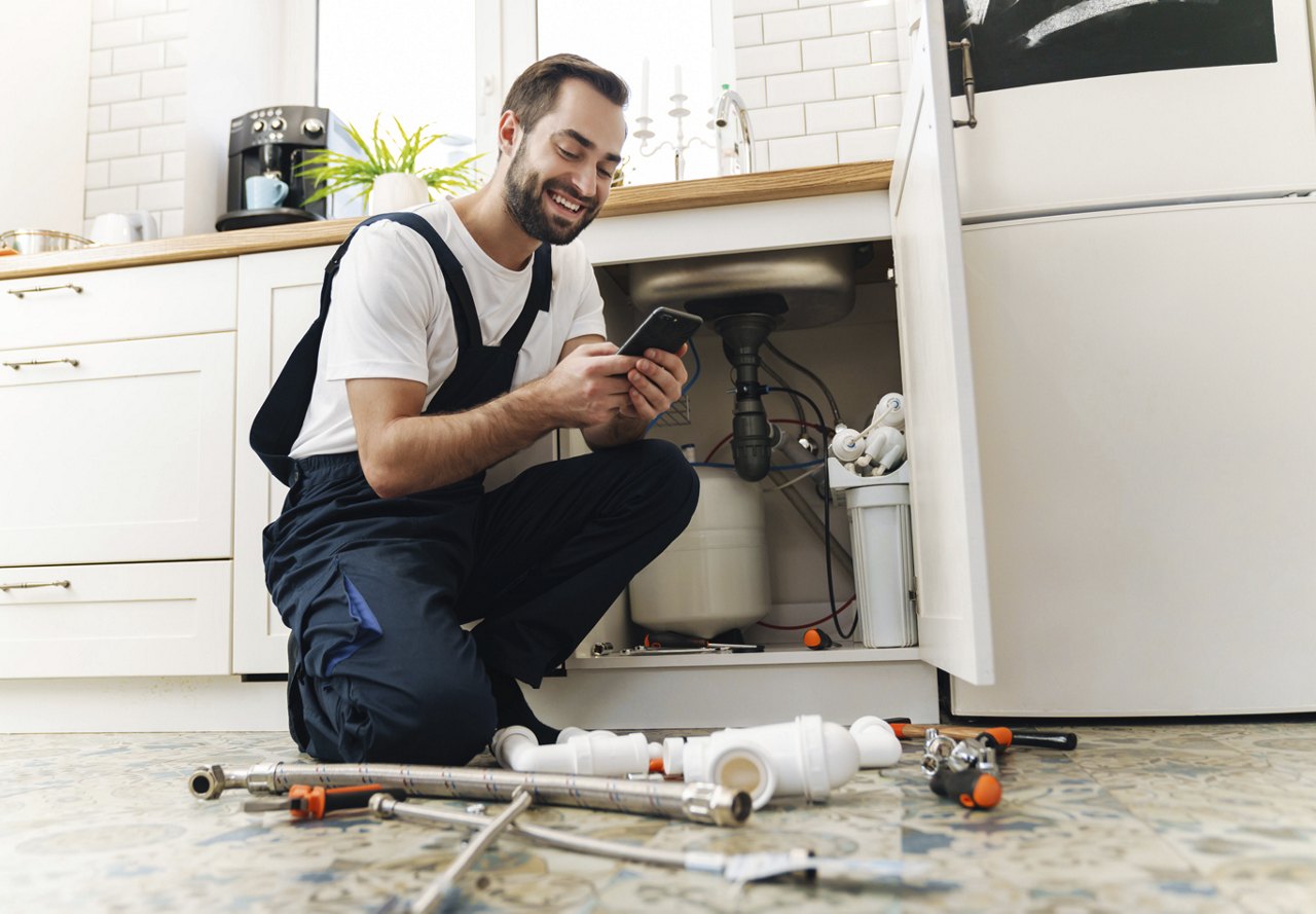 A maintenance worker repairs a sink in an apartment kitchen while checking his phone for details from the maintenance request.