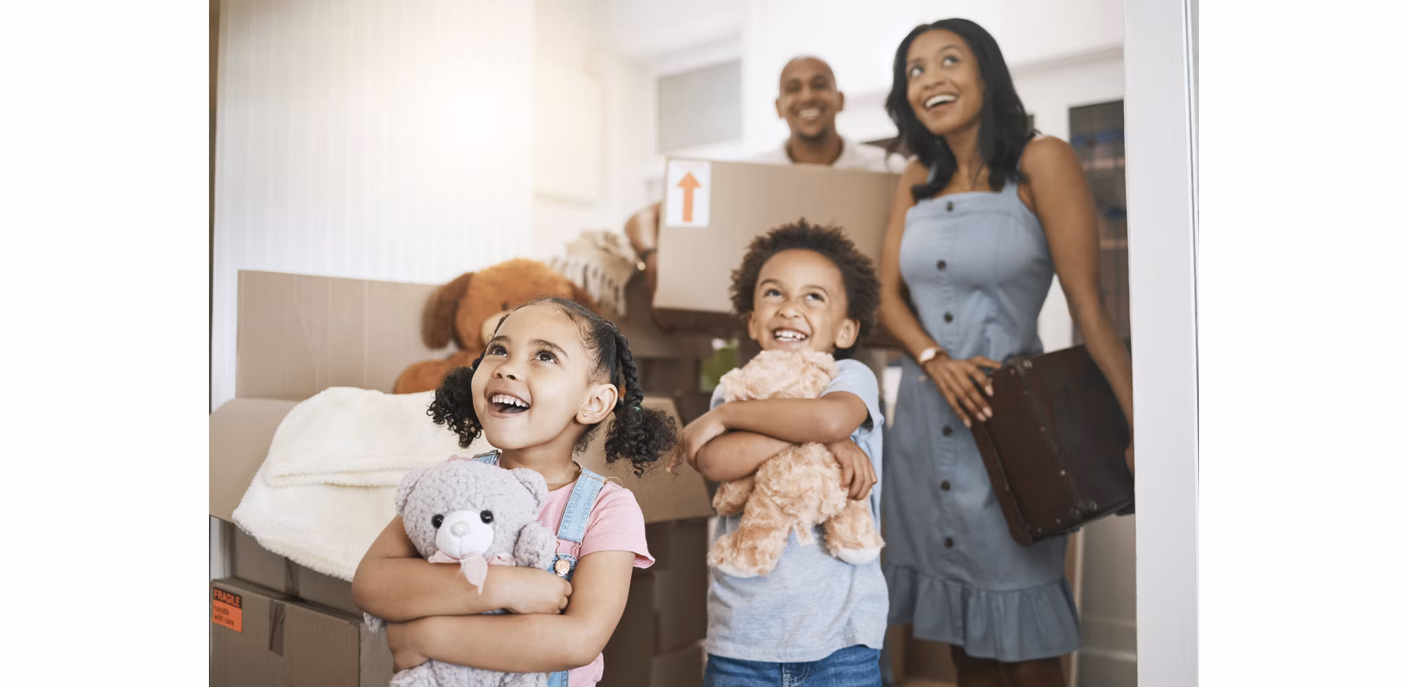 A family walking into their new home, with children holding stuffed animals and cardboard boxes, their faces filled with excitement and amazement as they take in their new surroundings.