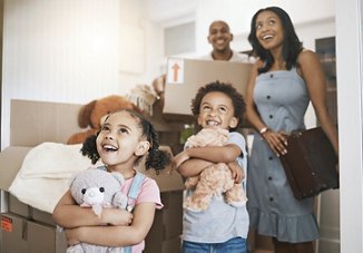 A family walking into their new home, with children holding stuffed animals and cardboard boxes, their faces filled with excitement and amazement as they take in their new surroundings.