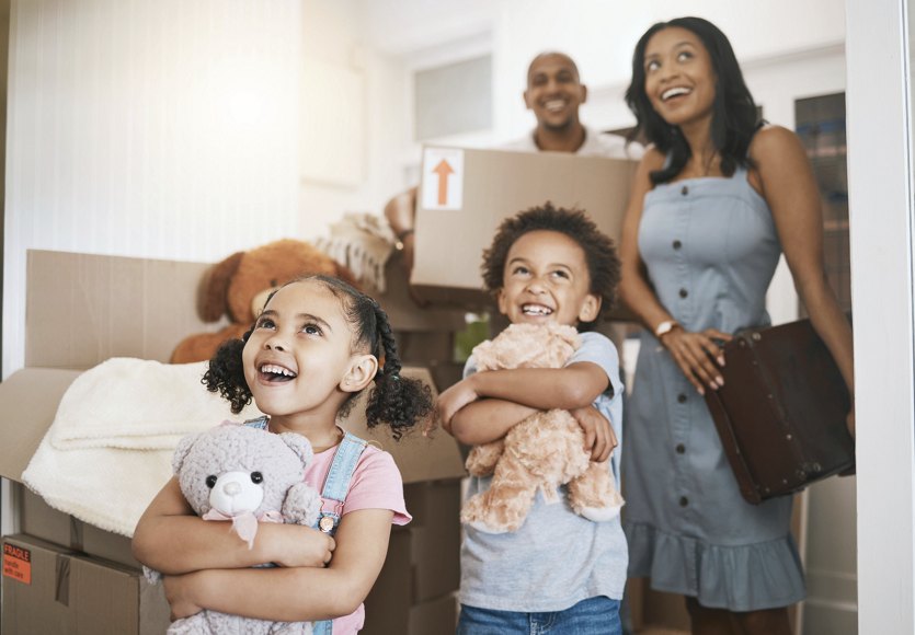 A family walking into their new home, with children holding stuffed animals and cardboard boxes, their faces filled with excitement and amazement as they take in their new surroundings.