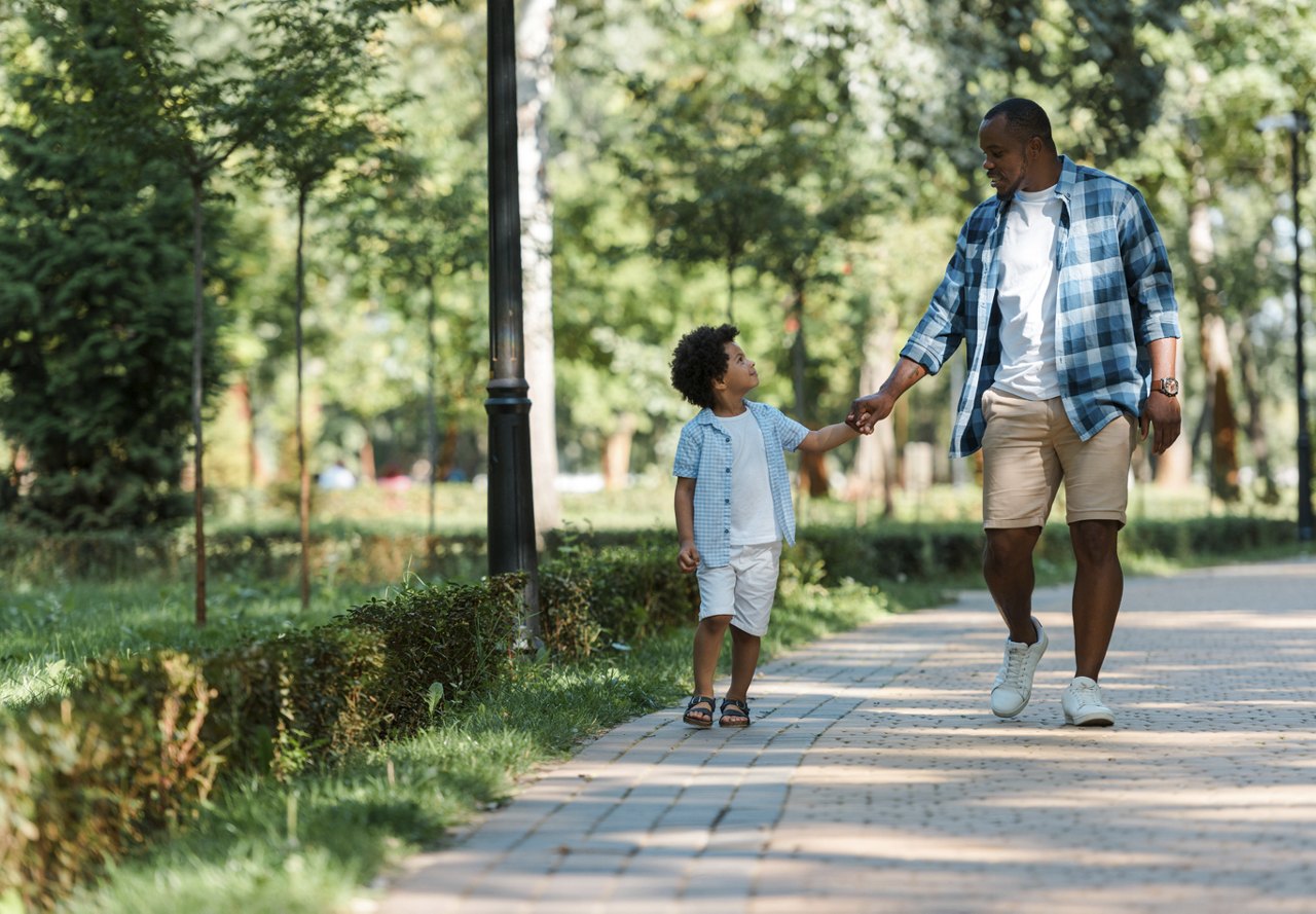 A father and his son walking through a sunlit park, surrounded by trees, sharing a warm and joyful moment as they look at each other.