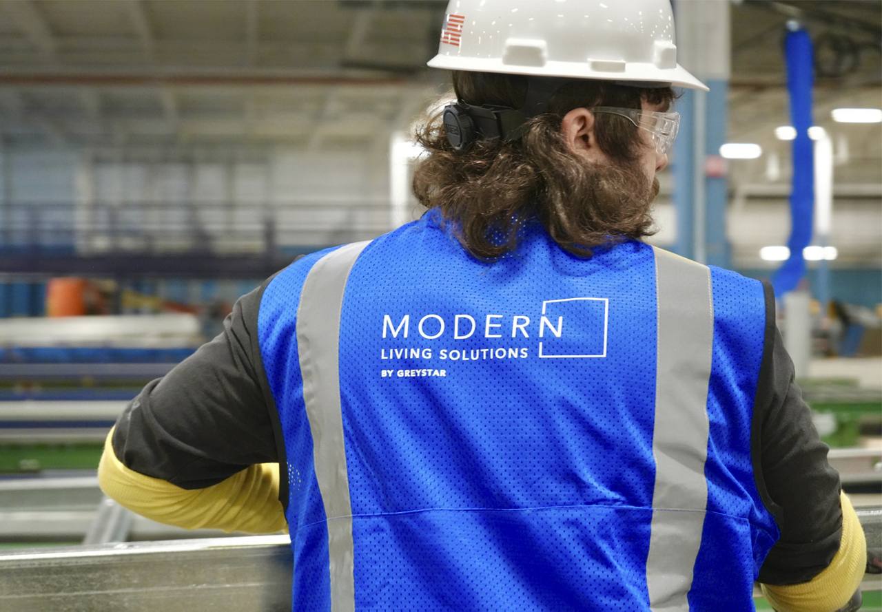 A worker in a blue 'MODERN Living Solutions' vest and safety helmet looking at industrial equipment.