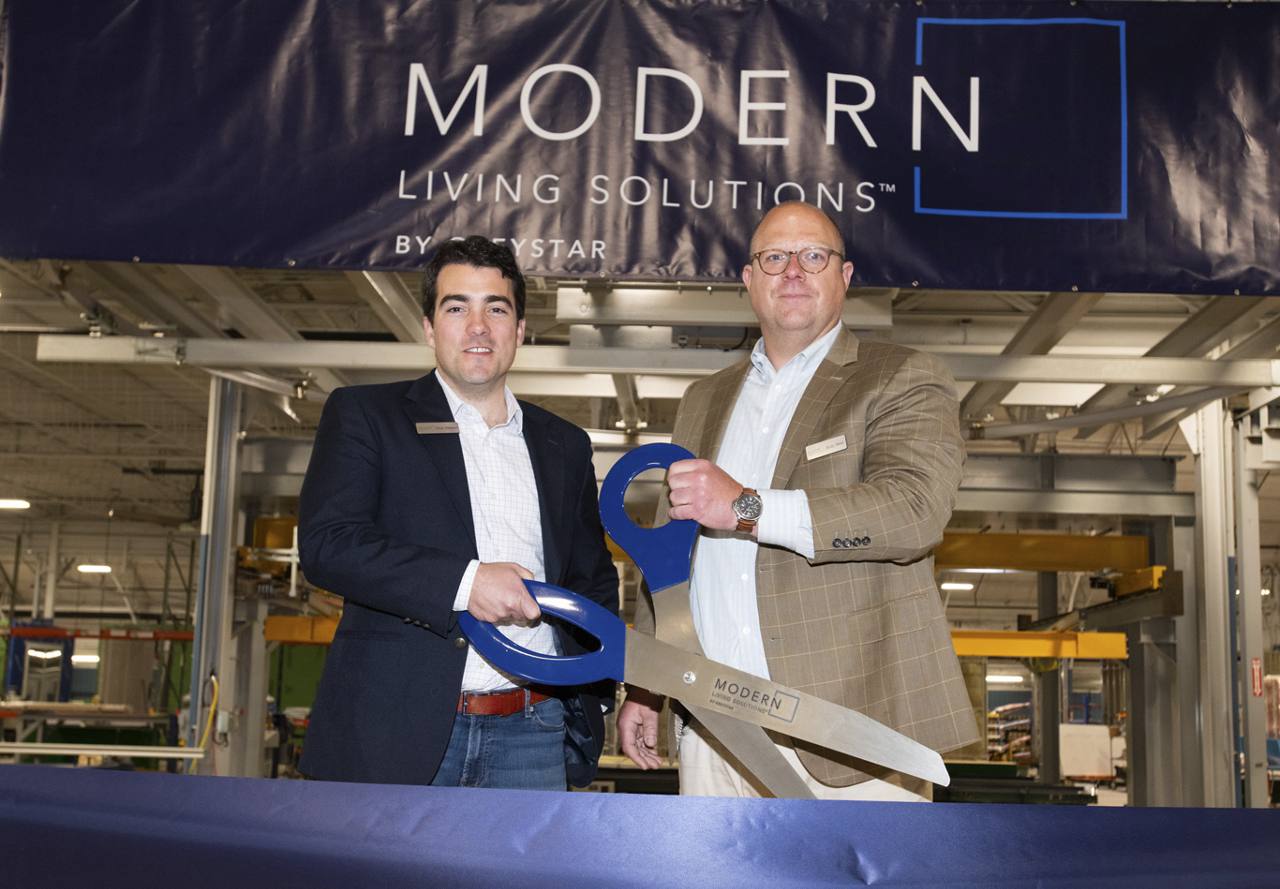 Two men in business attire standing in an industrial setting, smiling while holding a ceremonial ribbon in front of a banner that reads 'MODERN LIVING SOLUTIONS'.