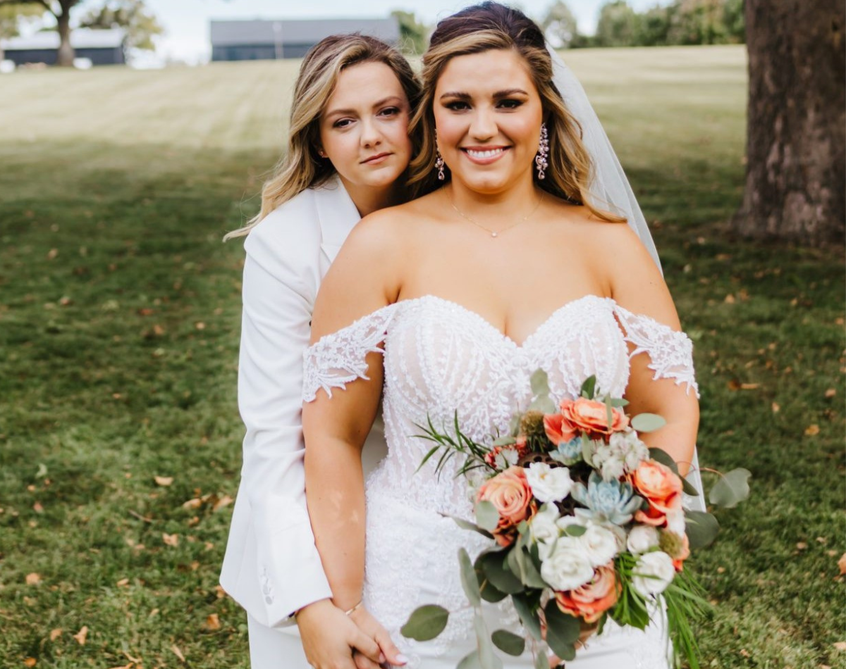 Two brides on their wedding day, one in a white suit and the other in a lace bridal gown, holding a bouquet, sharing a heartfelt moment.
