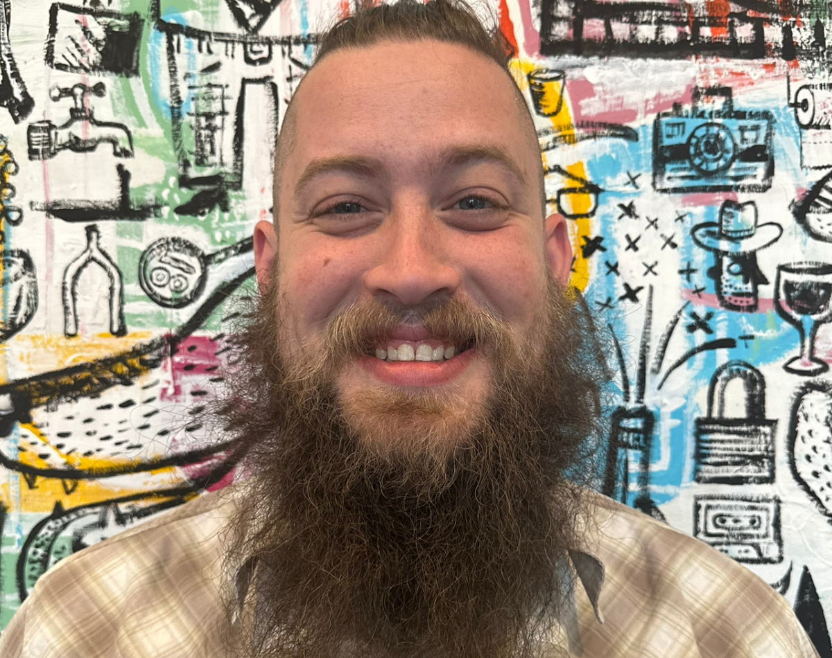 A bearded man with a wide smile in front of a colorful, graffiti-style mural.