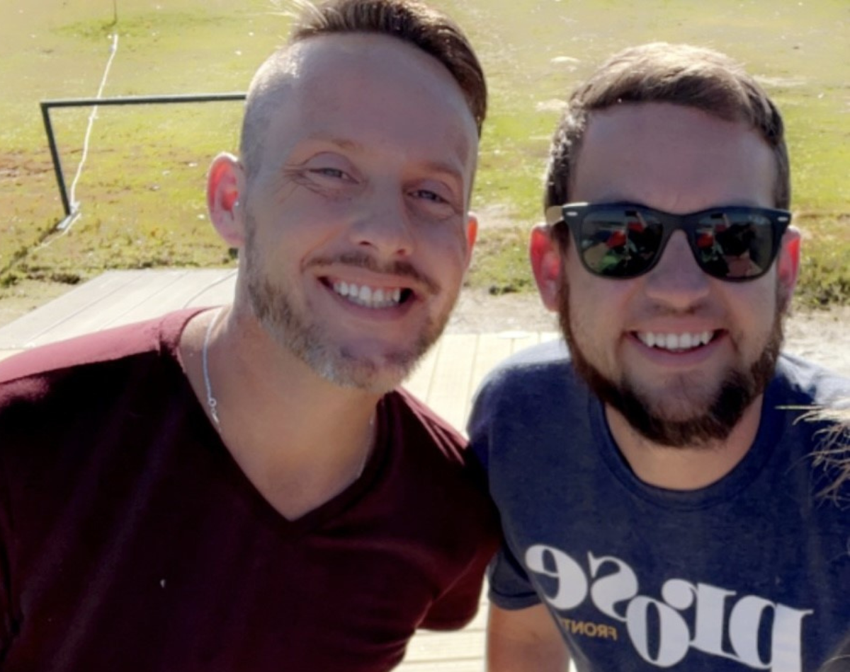 Two smiling men, one with a beard wearing sunglasses, outdoors in a sunny setting.