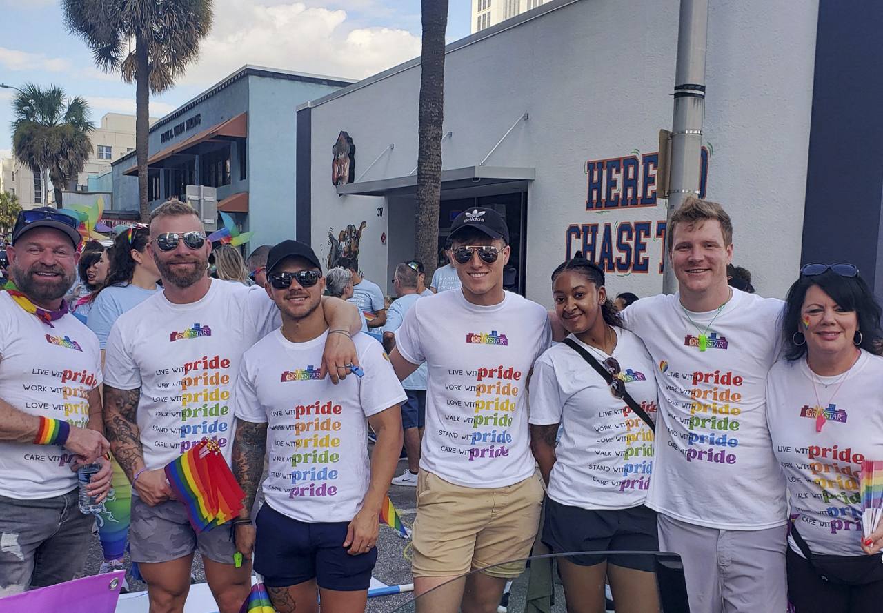 A happy group of individuals participating in a Pride event, wearing matching T-shirts with a rainbow-colored 'pride' slogan, and holding rainbow flags.