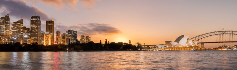 Twilight over a waterfront cityscape, featuring the iconic opera house and a distinctive bridge, with reflections on the water.