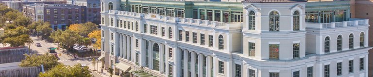 An aerial view of a stately white building with classic architecture, featuring prominent columns and arch windows. The building stands out amidst a cityscape of modern and traditional buildings, with a street running in front bordered by trees in autumn foliage.