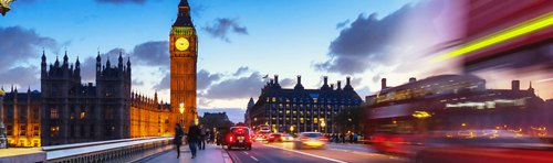 Evening traffic blurs past the illuminated Big Ben and the Houses of Parliament in London, under a dusky sky.
