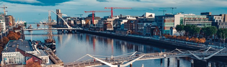 Scenic view of an Irish harbor with a canal, lined with a variety of buildings reflecting traditional and modern architectural styles. A charming bridge arches over the canal, adding to the picturesque urban landscape, under a clear sky.