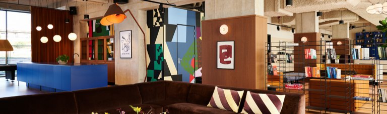 Modern cozy interior of a lounge with eclectic decor, featuring a large brown sofa, colorful abstract art, and a well-stocked bookshelf.