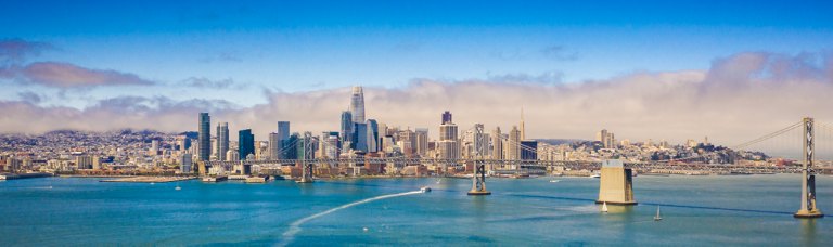 Panoramic view of a city skyline with skyscrapers and a famous suspension bridge spanning a blue bay under a partly cloudy sky.