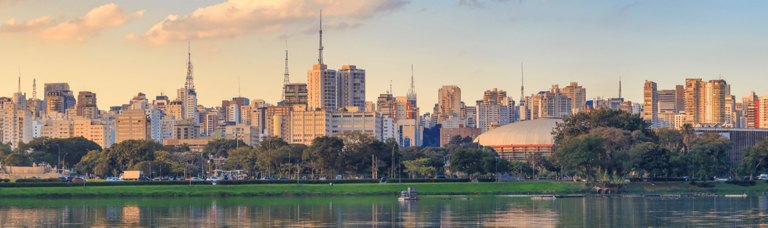 City skyline with dense high-rise buildings overlooking a calm lake with greenery in the foreground at sunset.