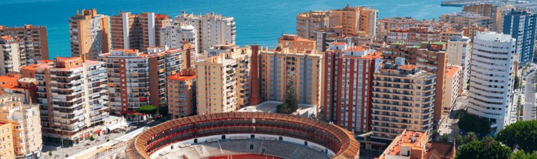 Aerial view of a dense urban area with colorful residential buildings and a large bullring, with the blue sea in the background.