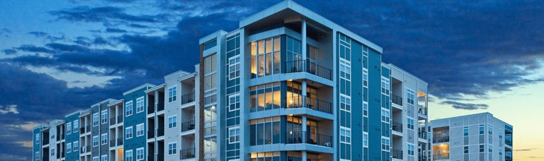 Captivating image of a modern apartment building in the United States at dusk, with its sleek, glass facade reflecting the stunning hues of the sunset sky. The building&apos;s angular corner provides a unique perspective, highlighting the blend of contemporary architecture and the natural beauty of the evening sky.