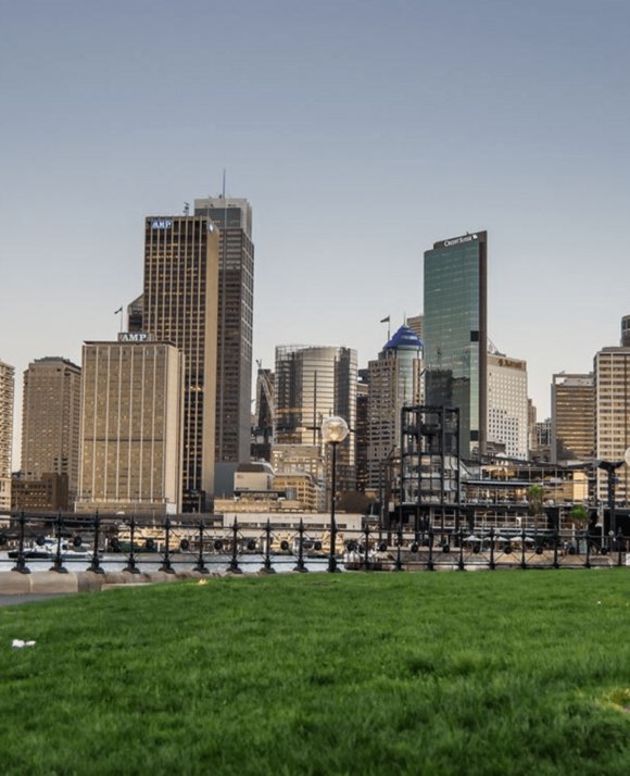 View of a city&apos;s high-rise buildings from a park with green grass in the foreground and a pedestrian bridge over a river.