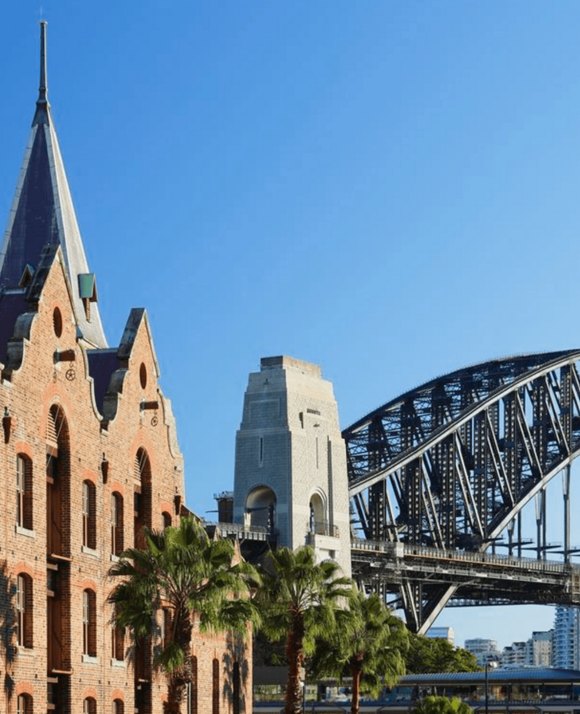 A historic stone building with a spire, palm trees in the foreground, and part of a large steel bridge structure visible in the background against a clear blue sky.