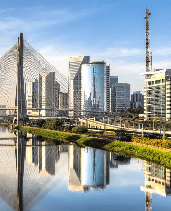 The Estaiada Bridge and its reflection on the river, with a backdrop of modern buildings under a clear sky in São Paulo, Brazil.