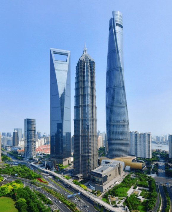 Aerial view of a cityscape with a trio of towering skyscrapers, each with unique architecture, alongside urban roads and green spaces.