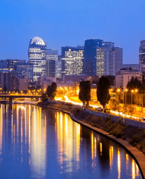 Twilight cityscape with a riverfront view, illuminated office buildings reflecting on the water, and a distinctive dome-shaped skyscraper.