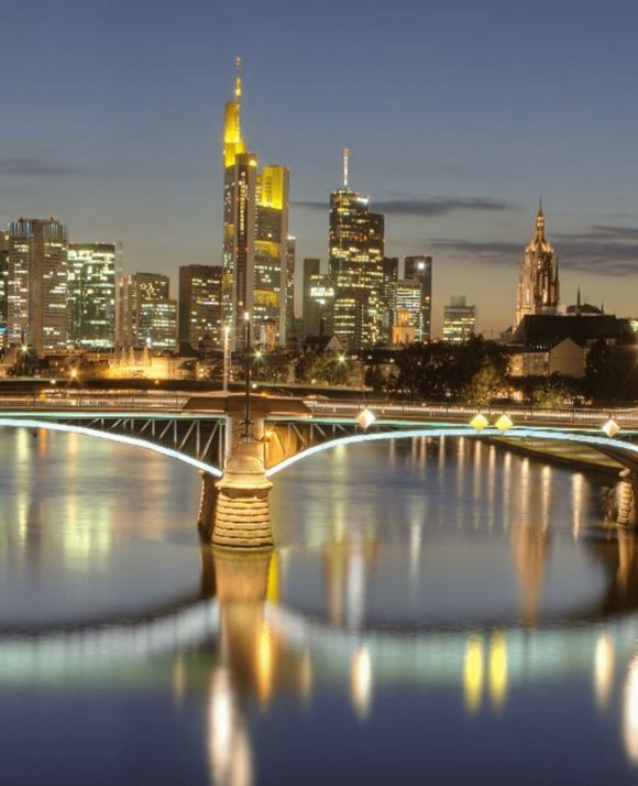Evening view of a city skyline with illuminated skyscrapers and a church, reflected in the calm waters of a river crossed by a bridge.