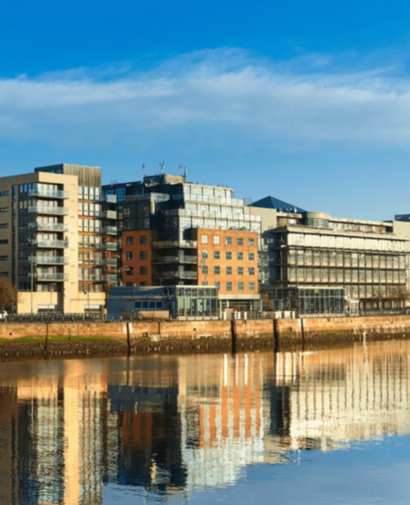 Modern waterfront buildings with reflective glass facades, casting their mirror image on the surface of a calm river under a blue sky.