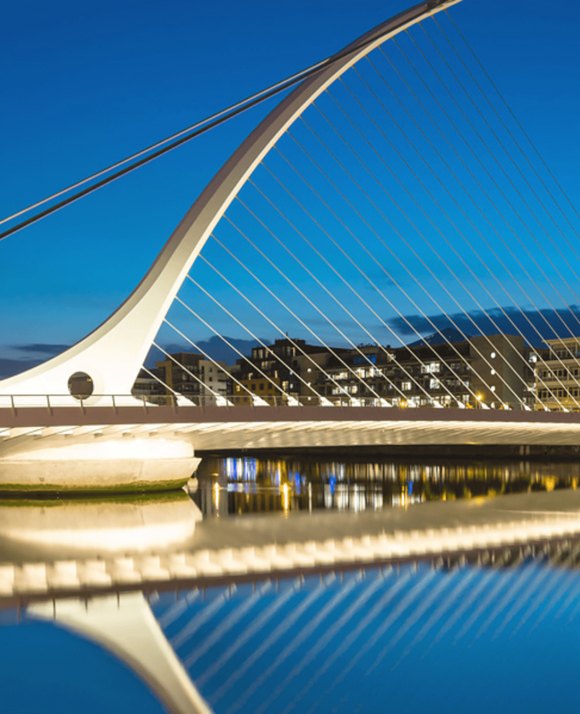 A modern cable-stayed bridge with a sweeping arch and reflective water surface at twilight, with city lights in the background.