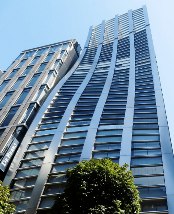 Upward view of a modern skyscraper with a sleek facade against a clear blue sky, framed by lush green foliage at the bottom.