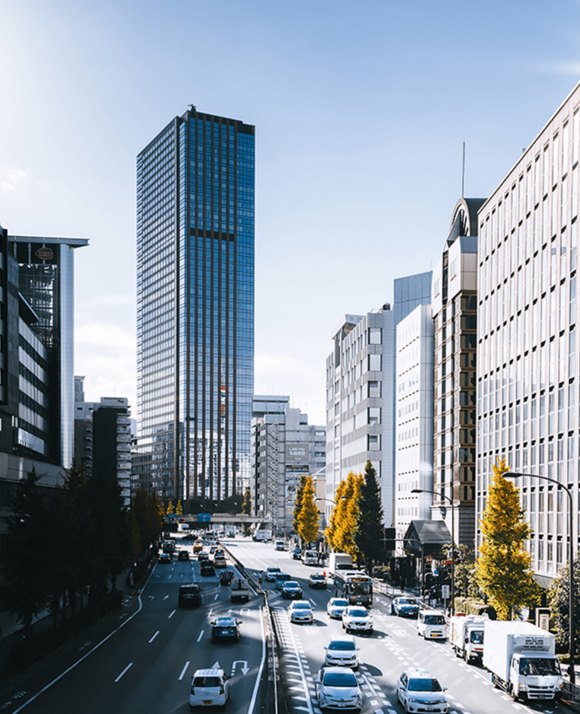 A bustling city street lined with tall office buildings under a clear blue sky, with autumn trees adding a splash of color.