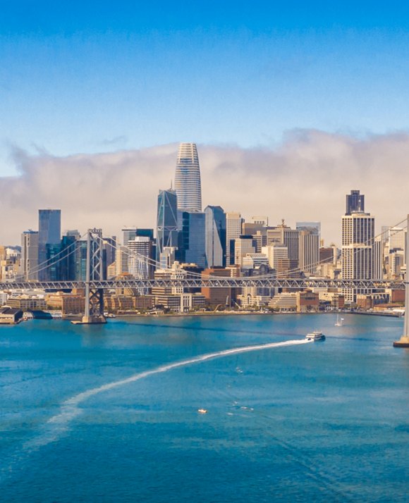 A panoramic view of a bustling city skyline with a prominent bridge over a blue body of water, under a clear sky.