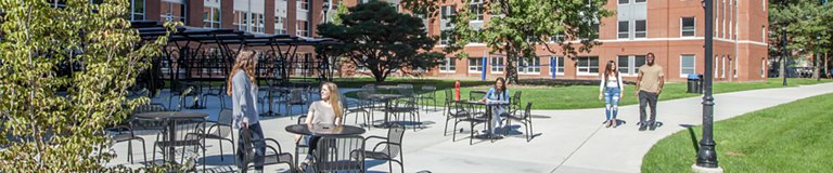 Students engage in various activities in an outdoor patio area with tables and chairs, set against a backdrop of campus buildings and greenery.