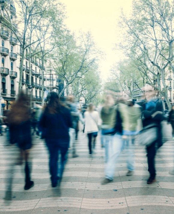 Blurry motion of people bustling down a tree-lined urban street with classic architecture, conveying the dynamic energy of city life.