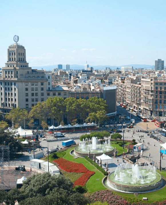 Aerial view of a bustling city square with ornate fountains, vibrant flower beds, and classic European architecture under a clear blue sky.