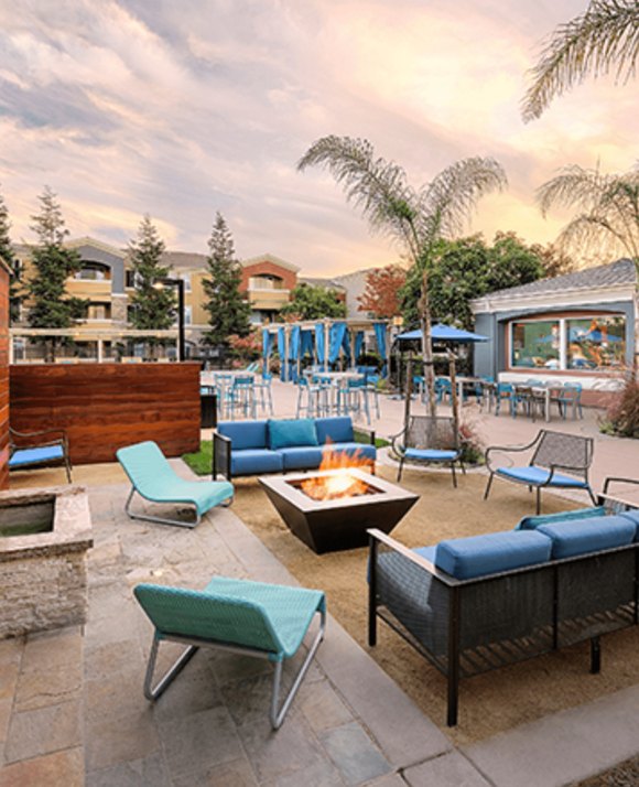 Outdoor residential lounge area with fire pit, blue seating, and palm trees under a cloudy sky.