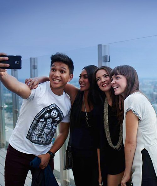 Four friends take a selfie together, smiling and huddled close, with a hazy city skyline in the background.