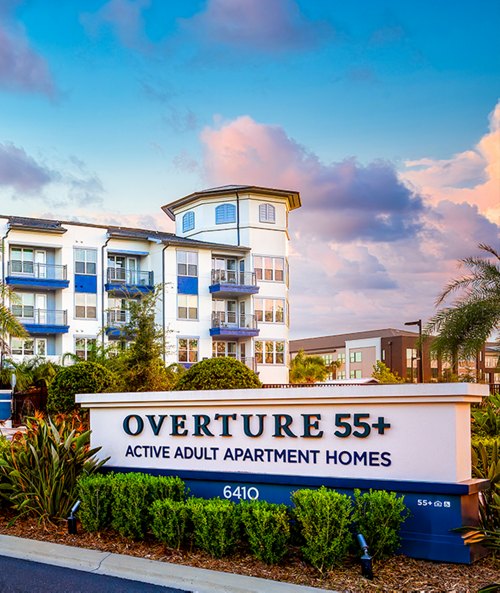 Entrance of Overture 55+ Active Adult Apartment Homes with a signboard in the foreground and a multi-story residential building in the background at dusk.