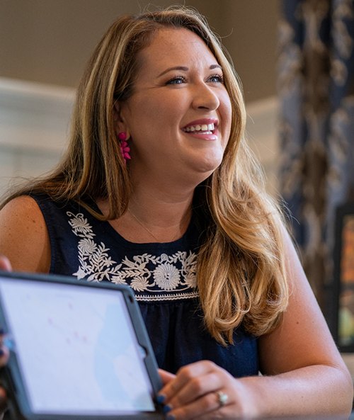 Smiling woman with blonde hair wearing a navy dress and pink earrings, holding a tablet, seemingly engaged in a cheerful conversation.