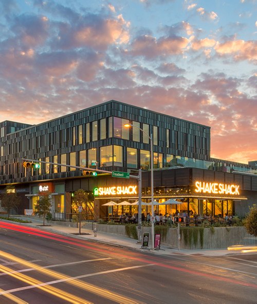 Exterior view of a modern Shake Shack restaurant at twilight with illuminated signage and visible patrons dining inside.