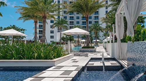 Resort-style pool walkway with black and white checkered tiles, surrounded by palm trees and white parasols.