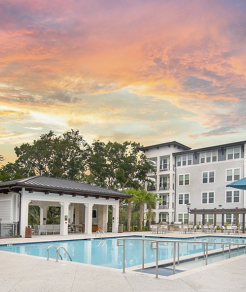 A tranquil outdoor pool area at dusk with a colorful sunset sky in hues of pink, orange, and blue. The pool is bordered by a covered pavilion with seating areas and the backdrop features a contemporary multi-story residential building with white and gray façade, surrounded by lush green trees.
