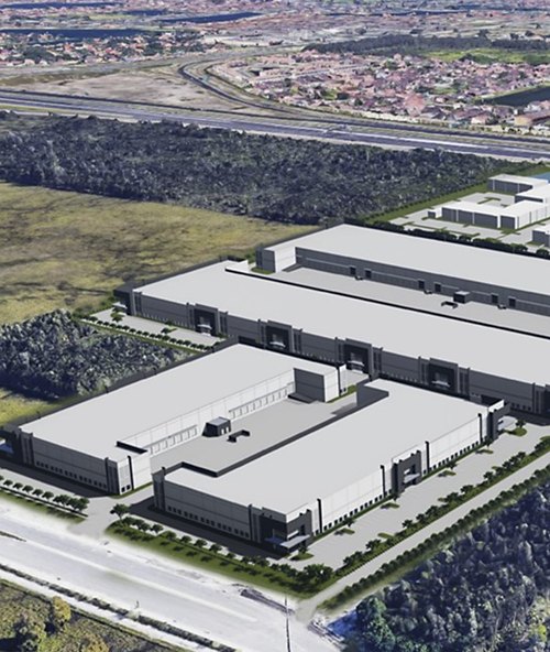 An aerial view of a large industrial complex with multiple warehouse buildings, surrounded by landscaping and a developing urban area in the background.