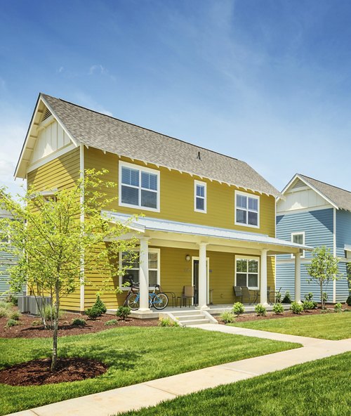 The image features a two-story yellow house with white trim and a covered porch, set in a well-landscaped yard under a blue sky. There's a blue bicycle parked in front of the house.