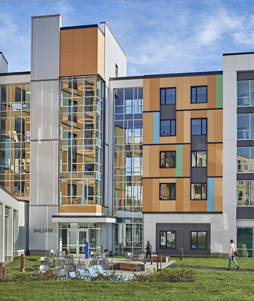 The photo showcases a modern student residence with a colorful facade and large windows. Outdoor communal spaces and passersby add life to the scene.