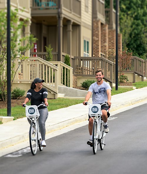 A man and a woman enjoy a leisurely bike ride together on a paved path in a residential area, with apartment buildings in the background.