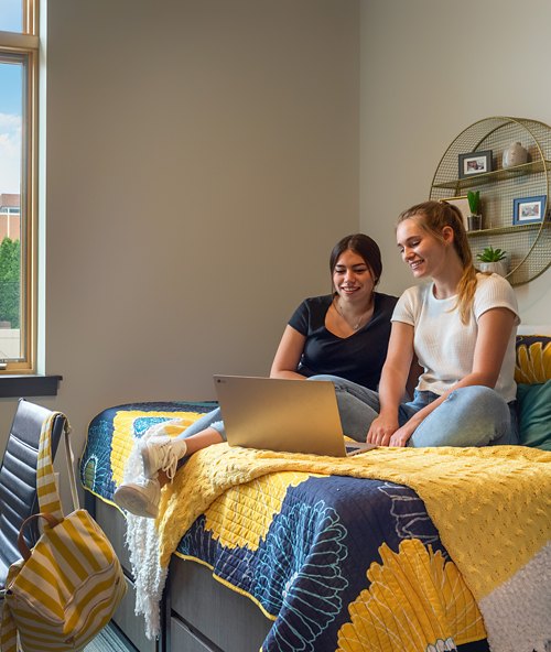 Two students are comfortably seated on a bed in a dorm room, sharing a moment of camaraderie over a laptop, possibly studying or enjoying leisure time together. The room exudes a warm, homey atmosphere with natural light streaming through the window.