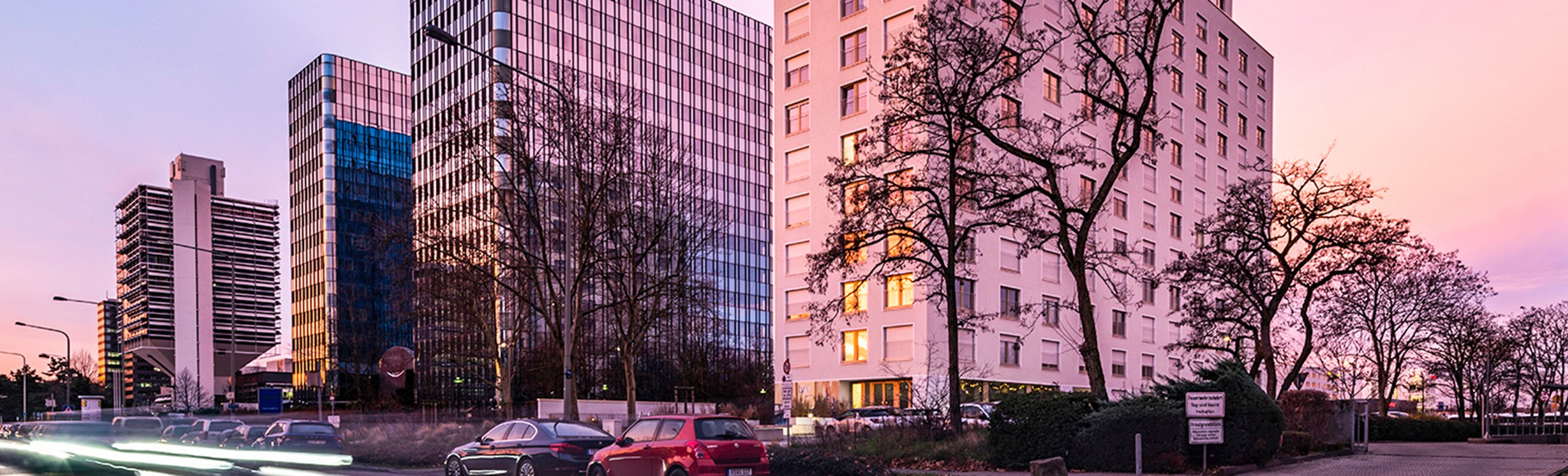 Panoramic view of a cityscape at dusk, showcasing modern high-rise buildings with reflective glass facades and a pastel-colored sky in the background.