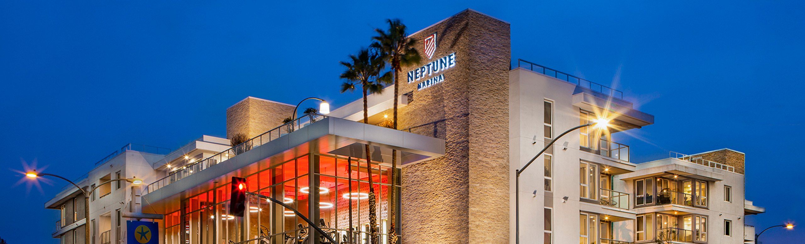 Twilight view of Neptune Marina residential building with illuminated windows and a clear sign, palm trees in the foreground under a dusk sky.